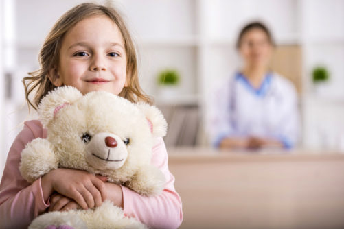 38975738 - little girl with teddy bear is looking at the camera. female doctor on background.
